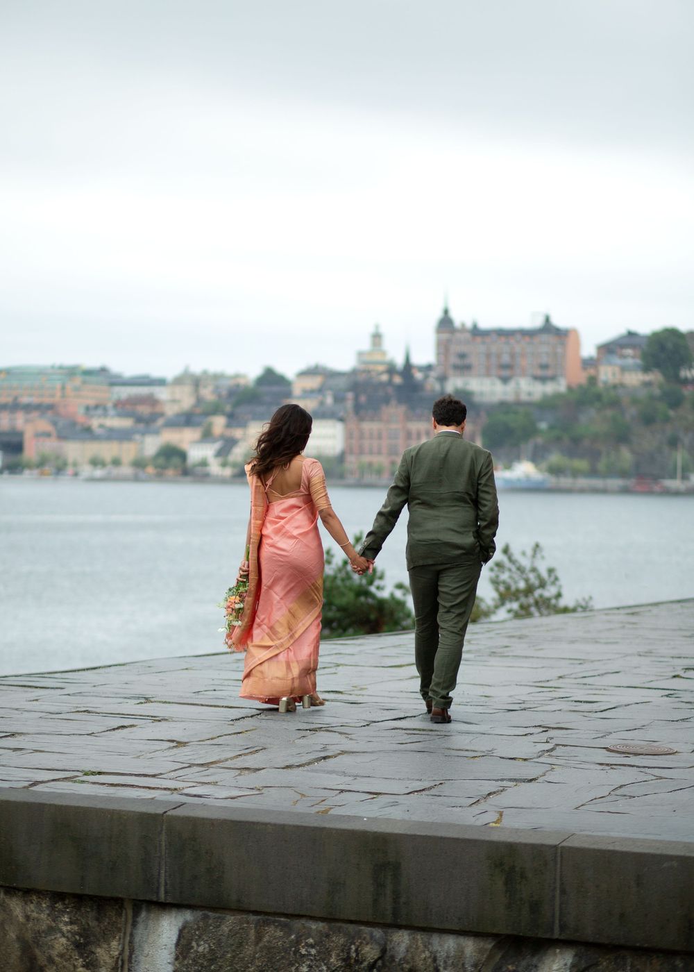 Couple in formal attire holding hands walking along a waterfront promenade with a historic cityscape in the background.
