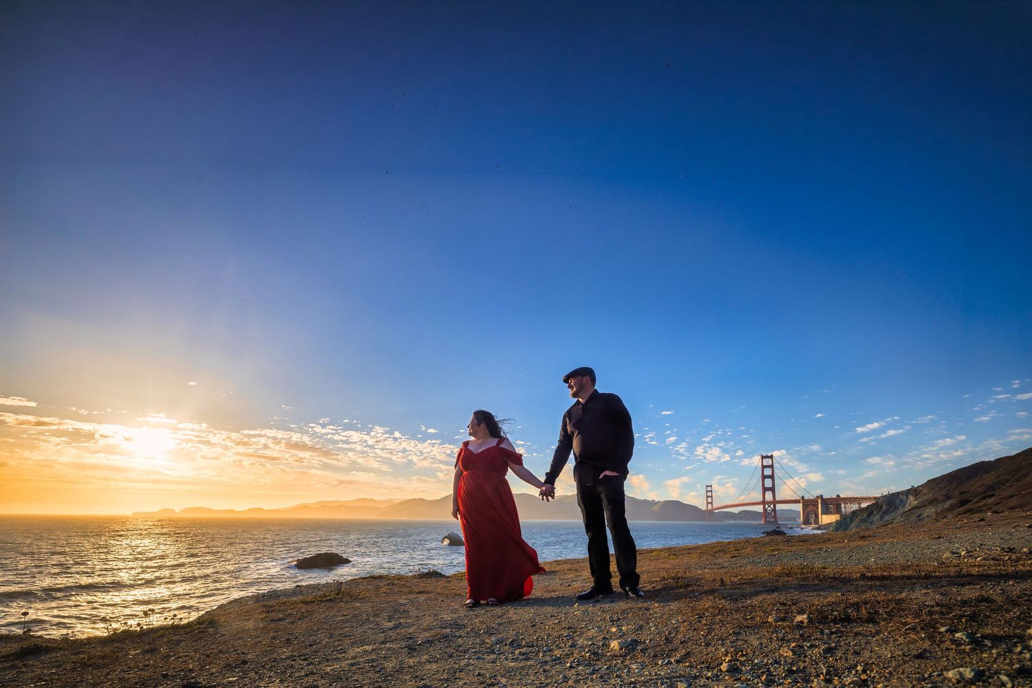 Couple holding hands at sunset on San Francisco beach with Golden Gate Bridge in background.