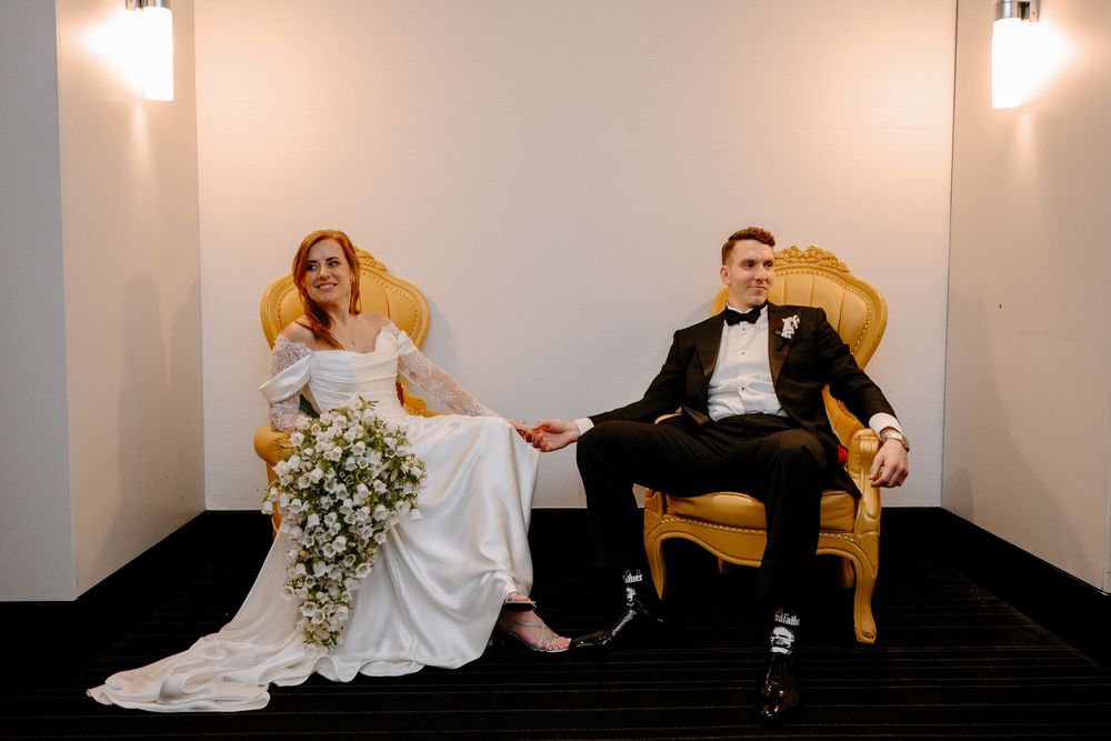 Bride and groom sitting in ornate gold chairs during wedding portrait session in elegant venue.