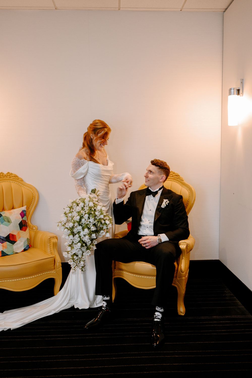 Bride in white gown holds grooms hand as he sits in a gold velvet chair during wedding portraits.