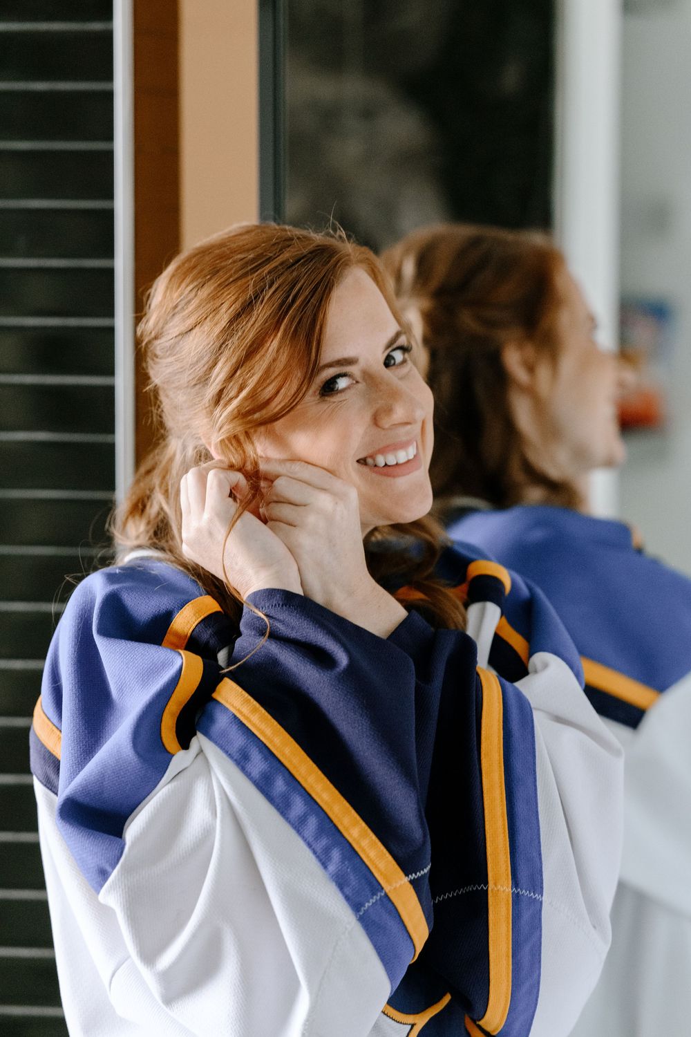 Smiling redhead woman in a blue and gold hockey jersey adjusting her hair near a mirror.
