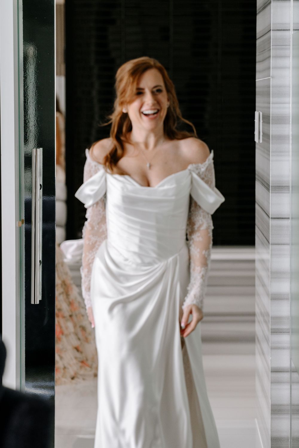 Joyful bride in an elegant off-shoulder white wedding gown with lace sleeves laughing indoors.