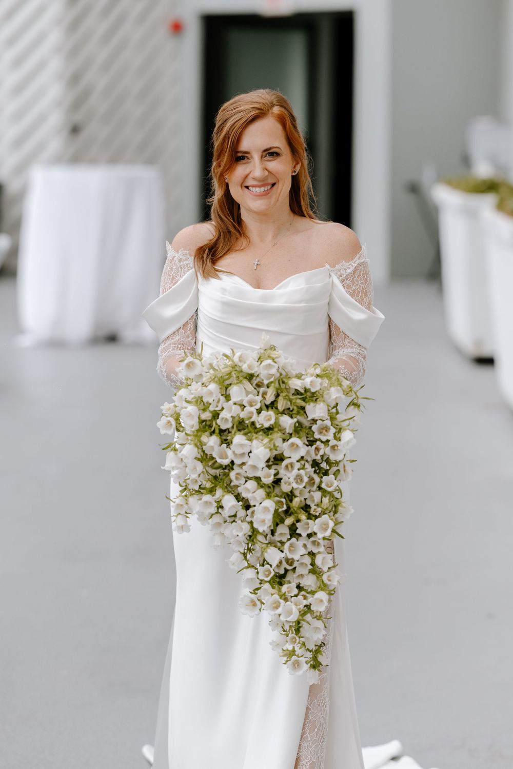 Smiling bride in off-shoulder white gown holding cascading bouquet of white roses and greenery indoors.