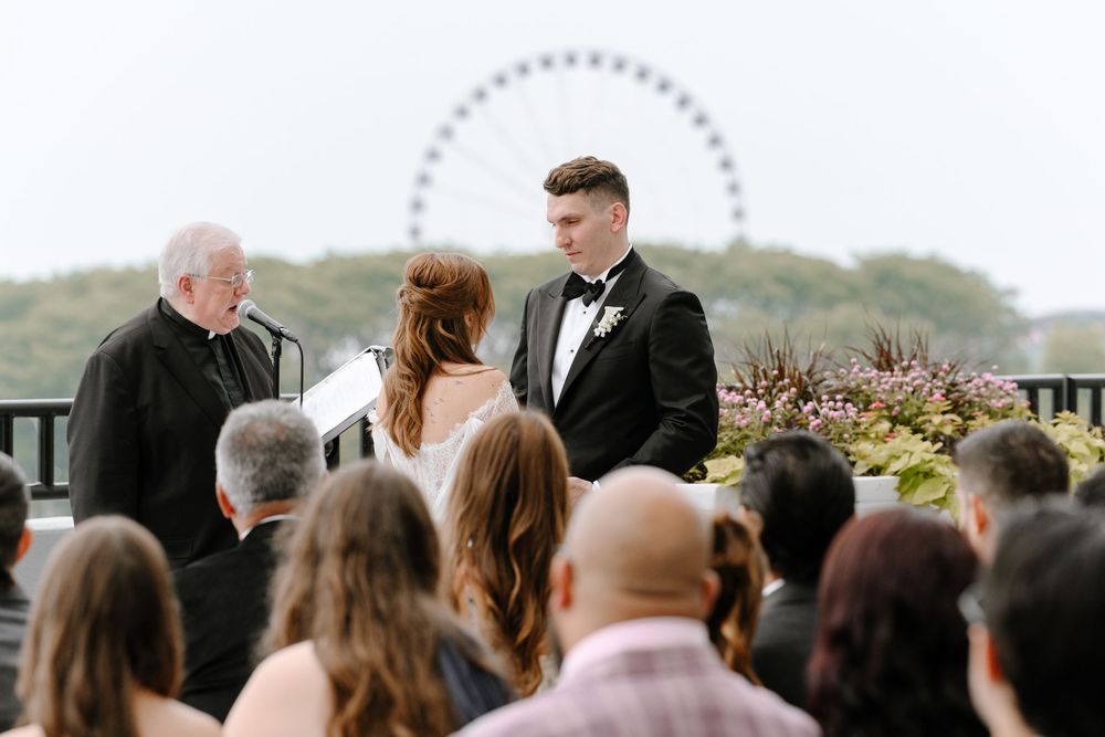 Outdoor rooftop wedding ceremony with couple exchanging vows, officiant, and Ferris wheel backdrop.