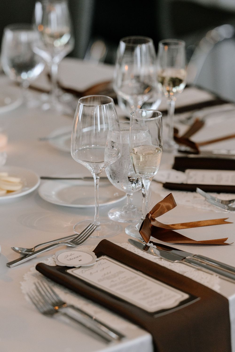 Elegant wedding reception table setting with crystal wine glasses, silver cutlery, brown napkins, and decorative menus on white linen.