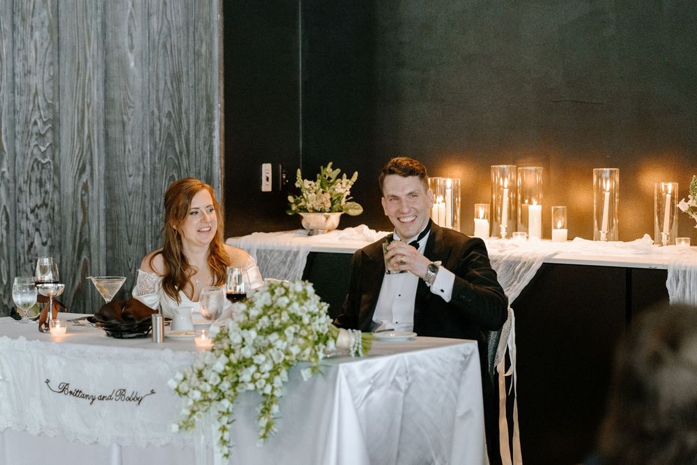 Smiling bride and groom seated at elegant sweetheart table with candles and white floral arrangements at wedding reception.