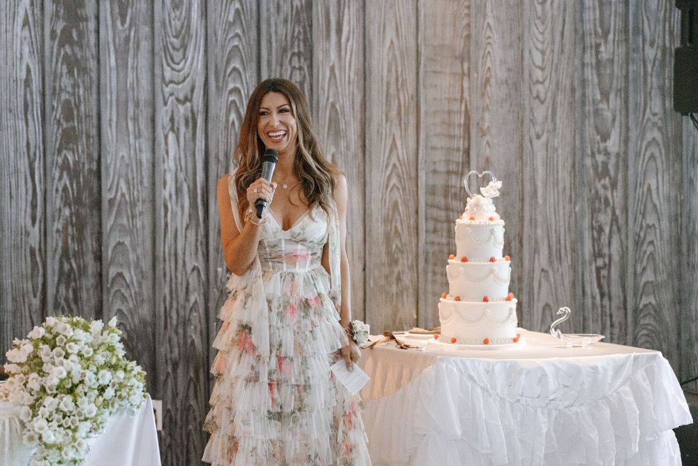 Woman in floral dress smiling holding microphone beside elegant tiered wedding cake at reception.