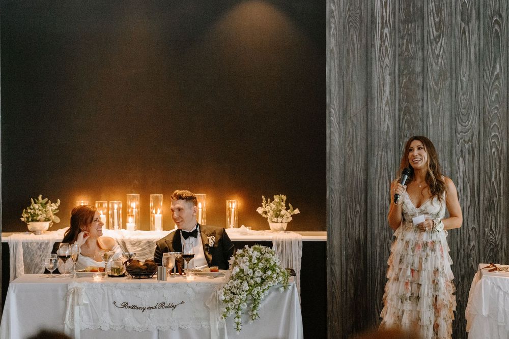 Woman in floral dress gives wedding speech at candlelit sweetheart table with bride and groom seated.