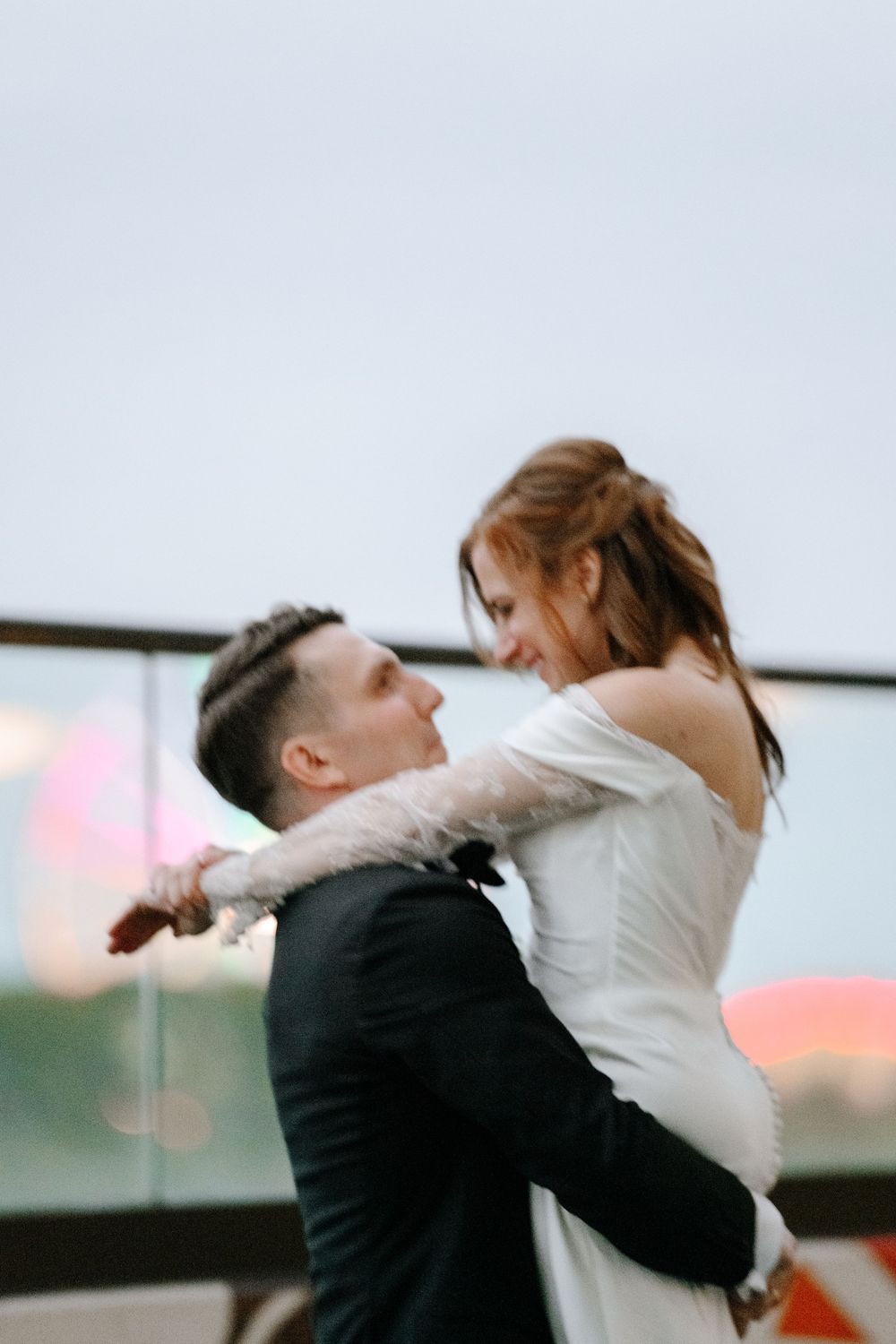 A groom in a black suit lifts his bride in a white off-shoulder dress, sharing a romantic moment outdoors.