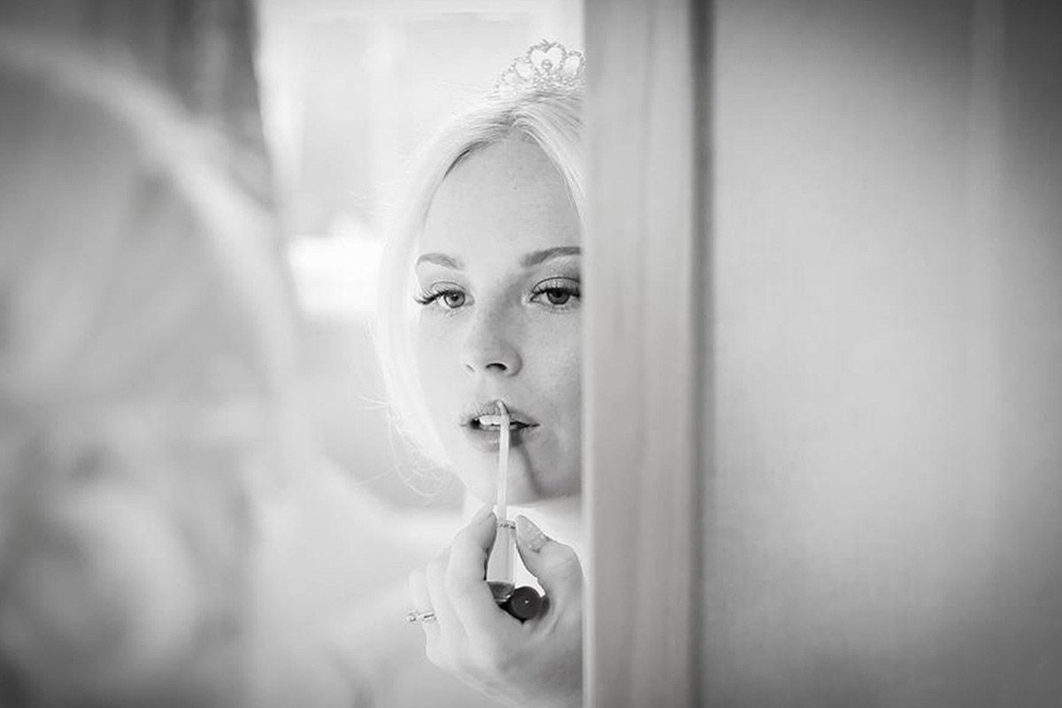 Black and white photo of a bride applying lipstick while looking in a mirror, wearing a tiara on her wedding day.