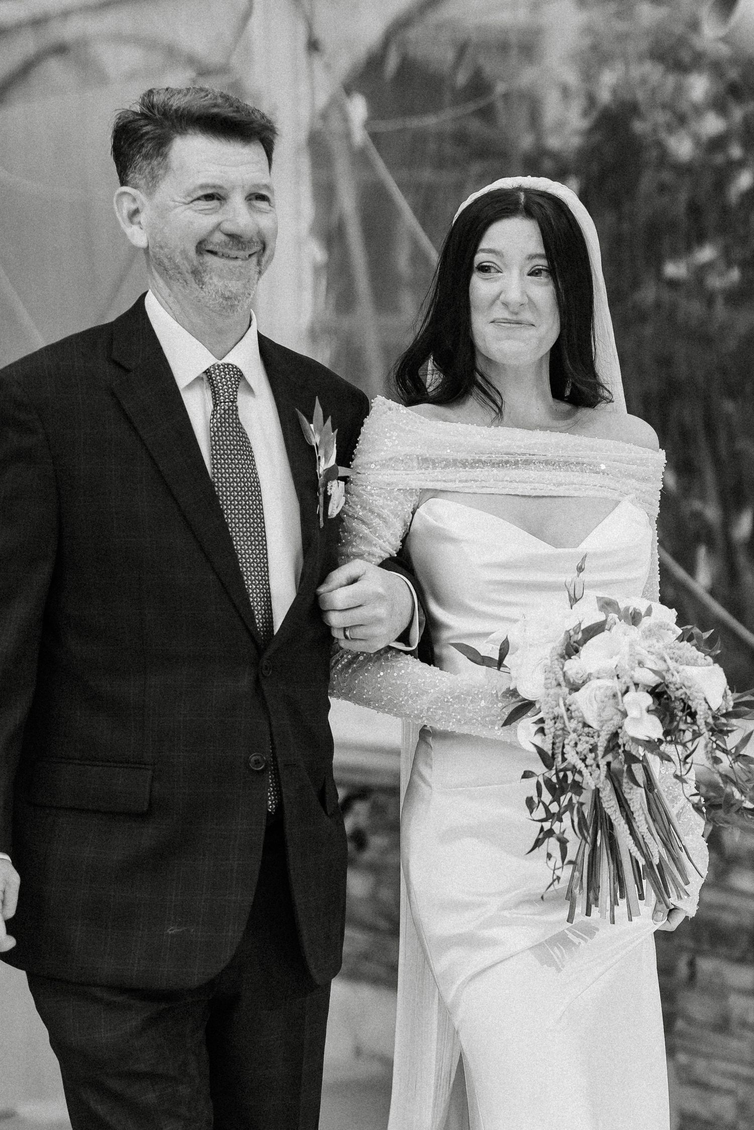 Black and white photo of a smiling groom in a suit walking arm in arm with a bride in a white gown holding a bouquet.
