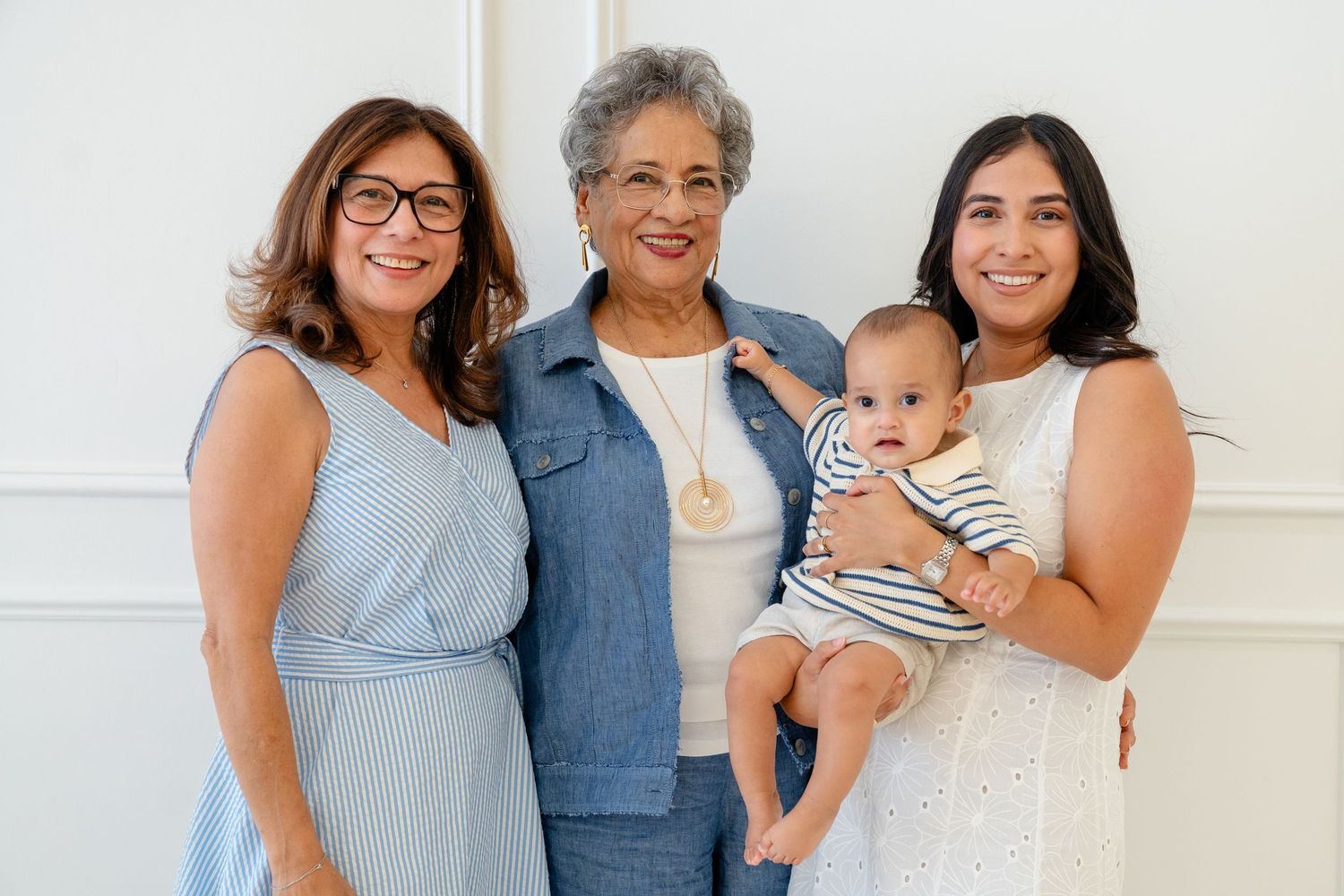 Three generations of women smile together, with the youngest holding a baby in a striped outfit, posing indoors.