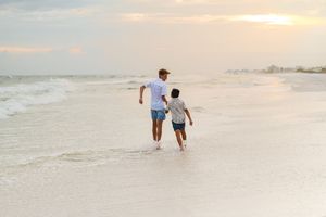 A father and child walk along a sandy beach at sunset, with gentle ocean waves washing over their feet.