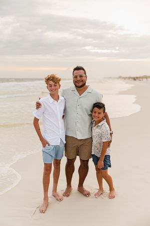 A man and two boys smile together on a sandy beach at sunset, wearing casual summer outfits with ocean waves behind them.