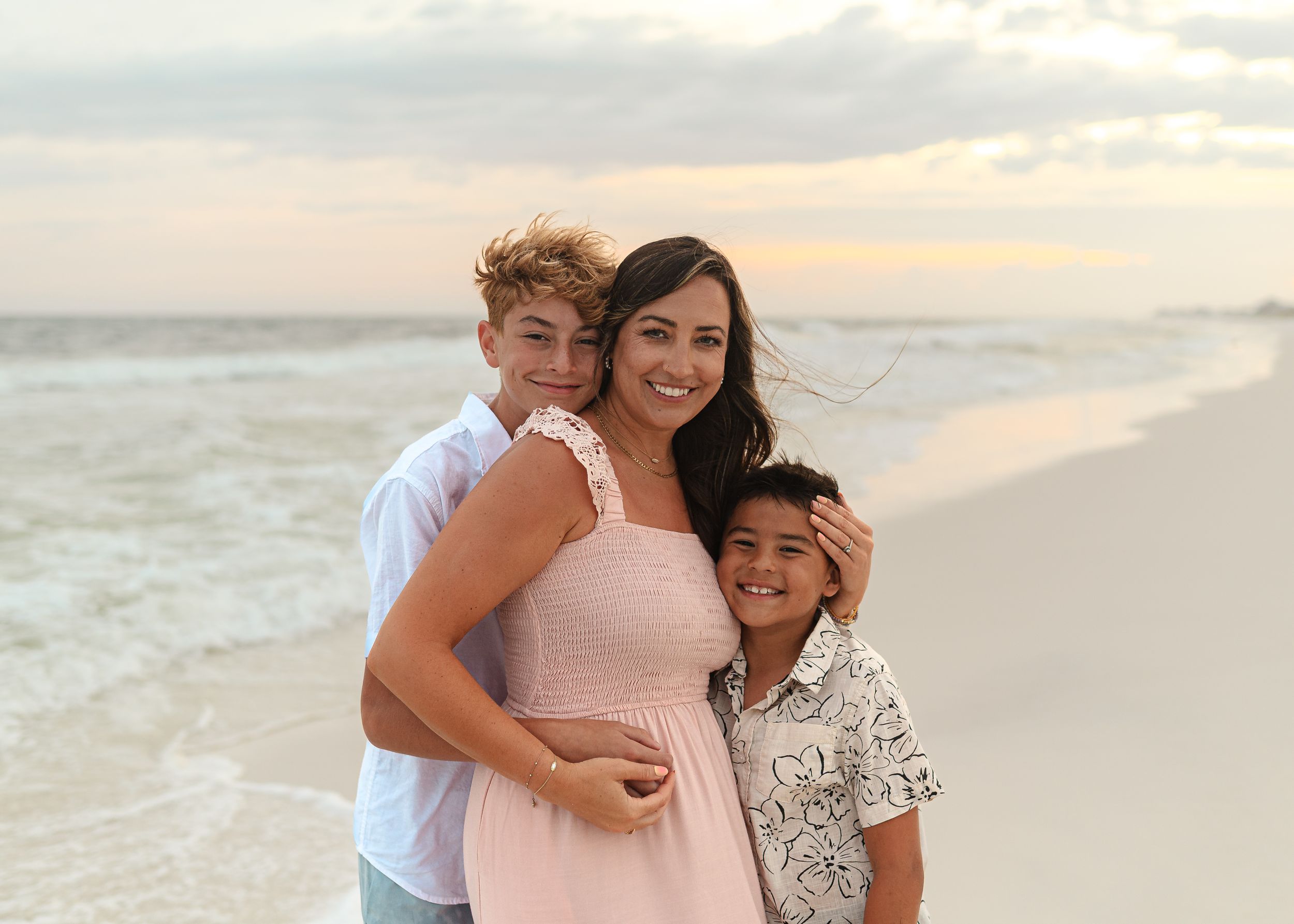 A woman and two boys smile together on a sandy beach at sunset in a warm family portrait.