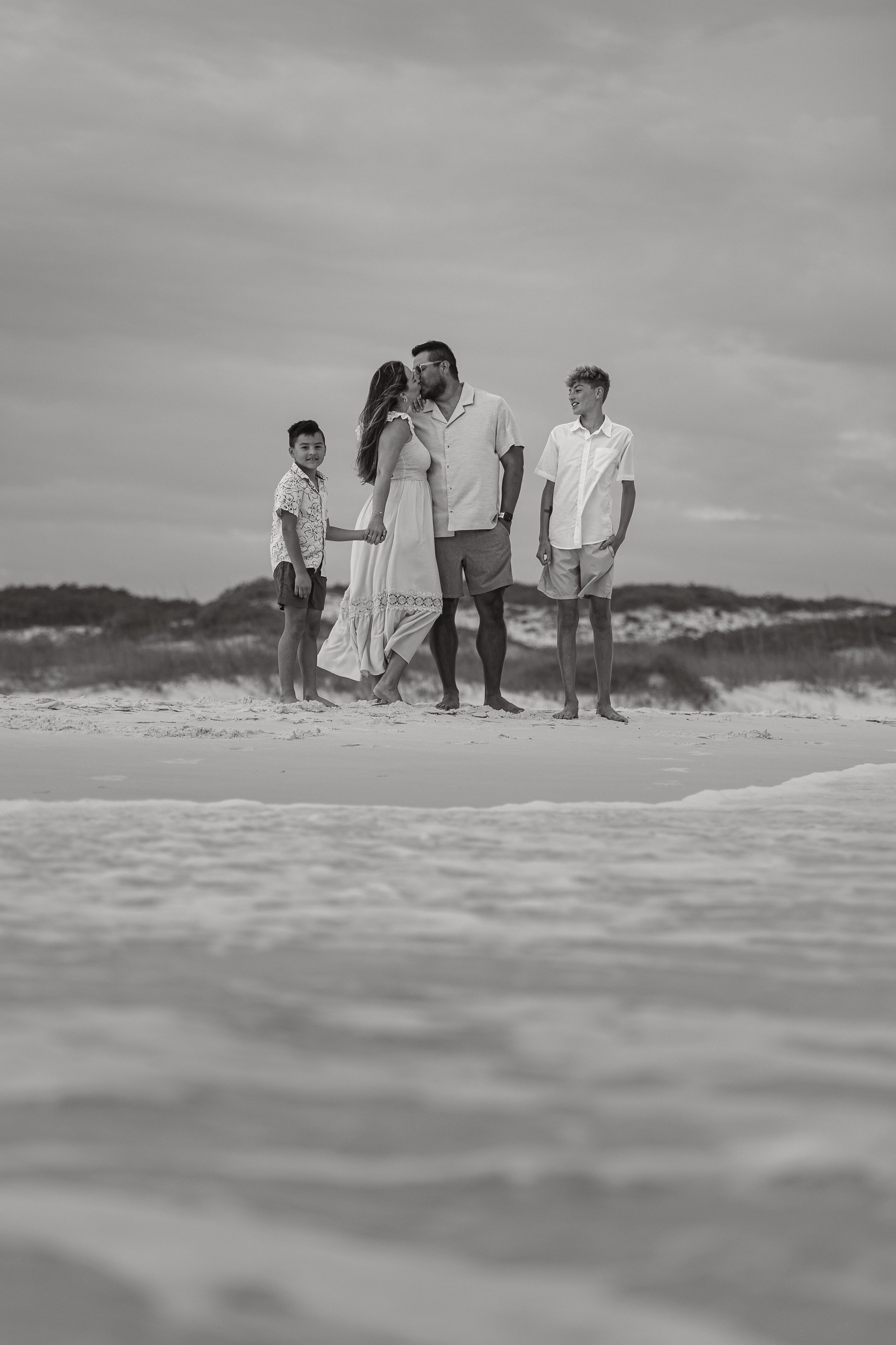 Black and white family portrait on beach with parents and two boys standing on sandy shore under cloudy sky.