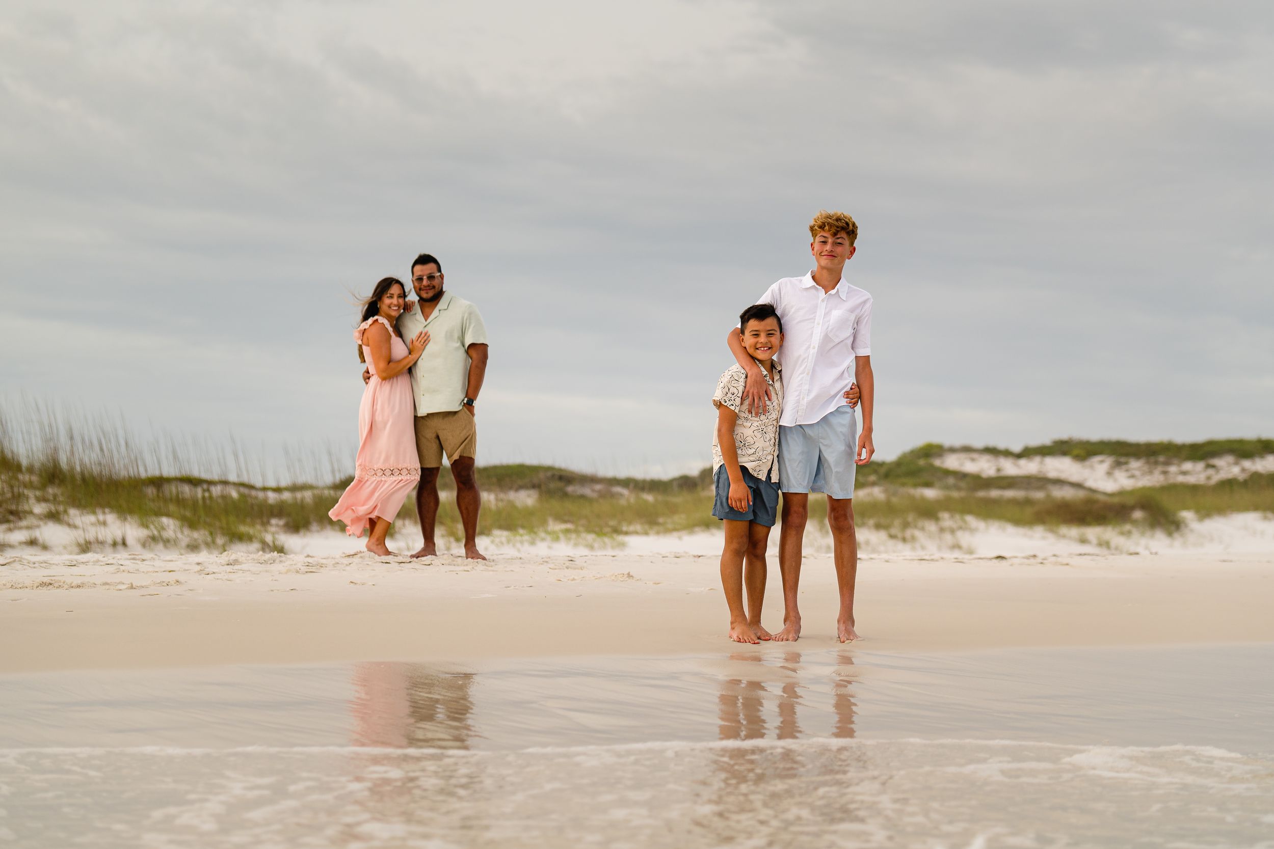 Family of four poses on a white sand beach with sea oats and ocean waves, two children in front, parents embracing behind.
