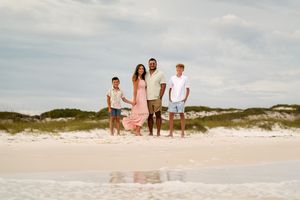 Family of four posing together on a white sandy beach with sand dunes and cloudy sky in the background.