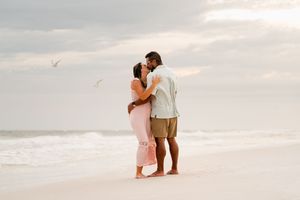 Couple sharing a romantic kiss on a sandy beach with ocean waves and cloudy sky in the background.