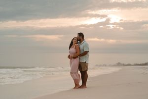 Couple embracing on a sandy beach at sunset, waves in background, woman in pink dress, man in casual attire.