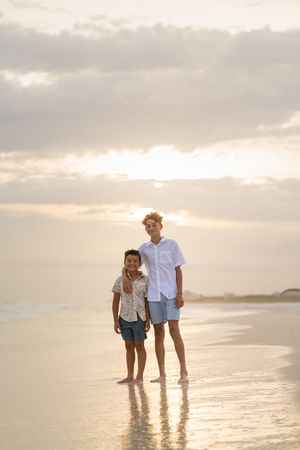 Two boys standing barefoot on a wet sandy beach at sunset, smiling at the camera with ocean waves behind them.