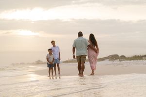Family of four walking barefoot on a sandy beach at sunset, with gentle waves and a golden sky.