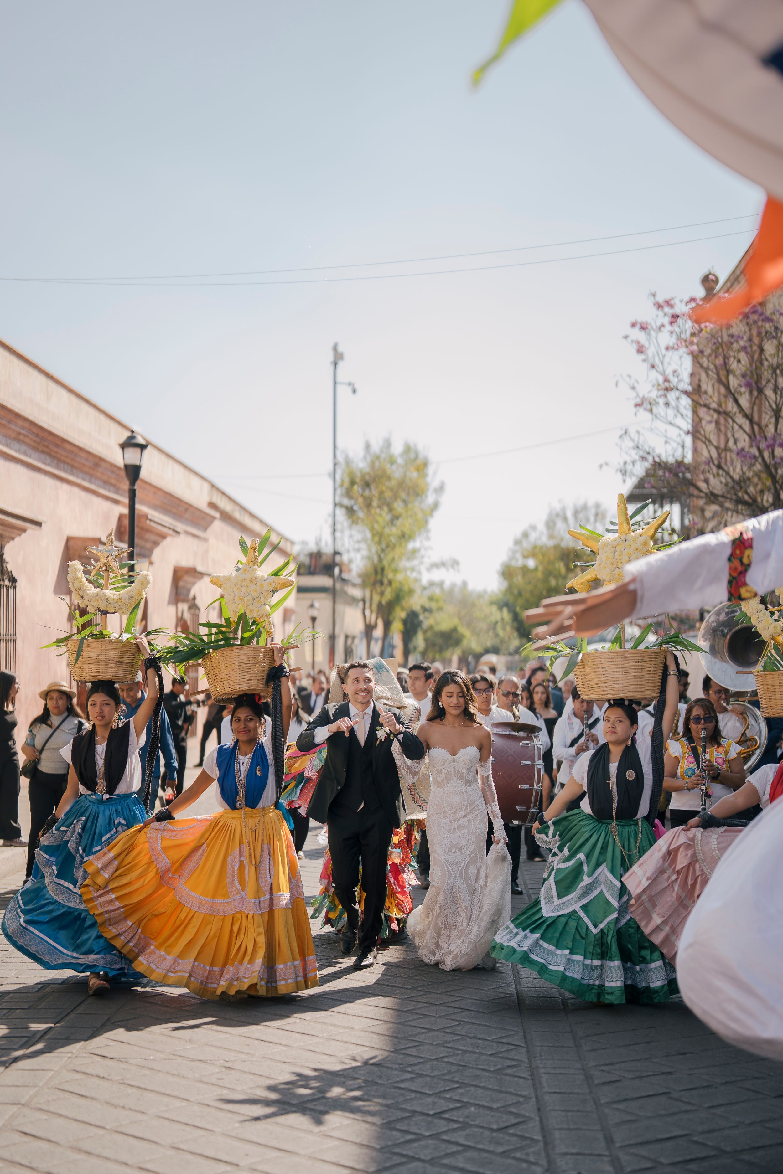 Bride and groom walk in colorful Mexican wedding parade with dancers in traditional dresses carrying baskets on their heads.