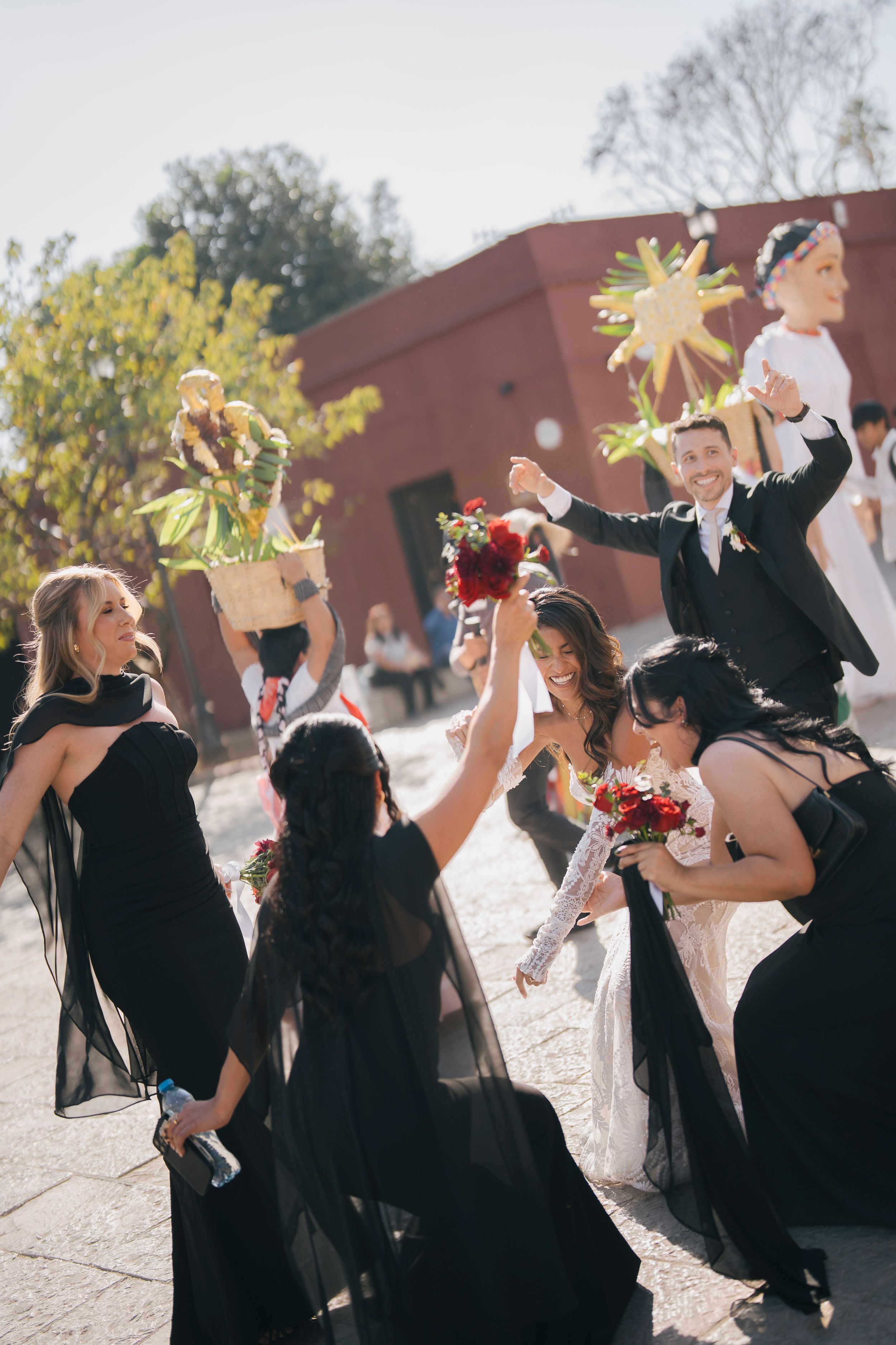 Wedding party in black dresses celebrating outdoors with bride and groom, holding colorful pinatas in sunny courtyard.