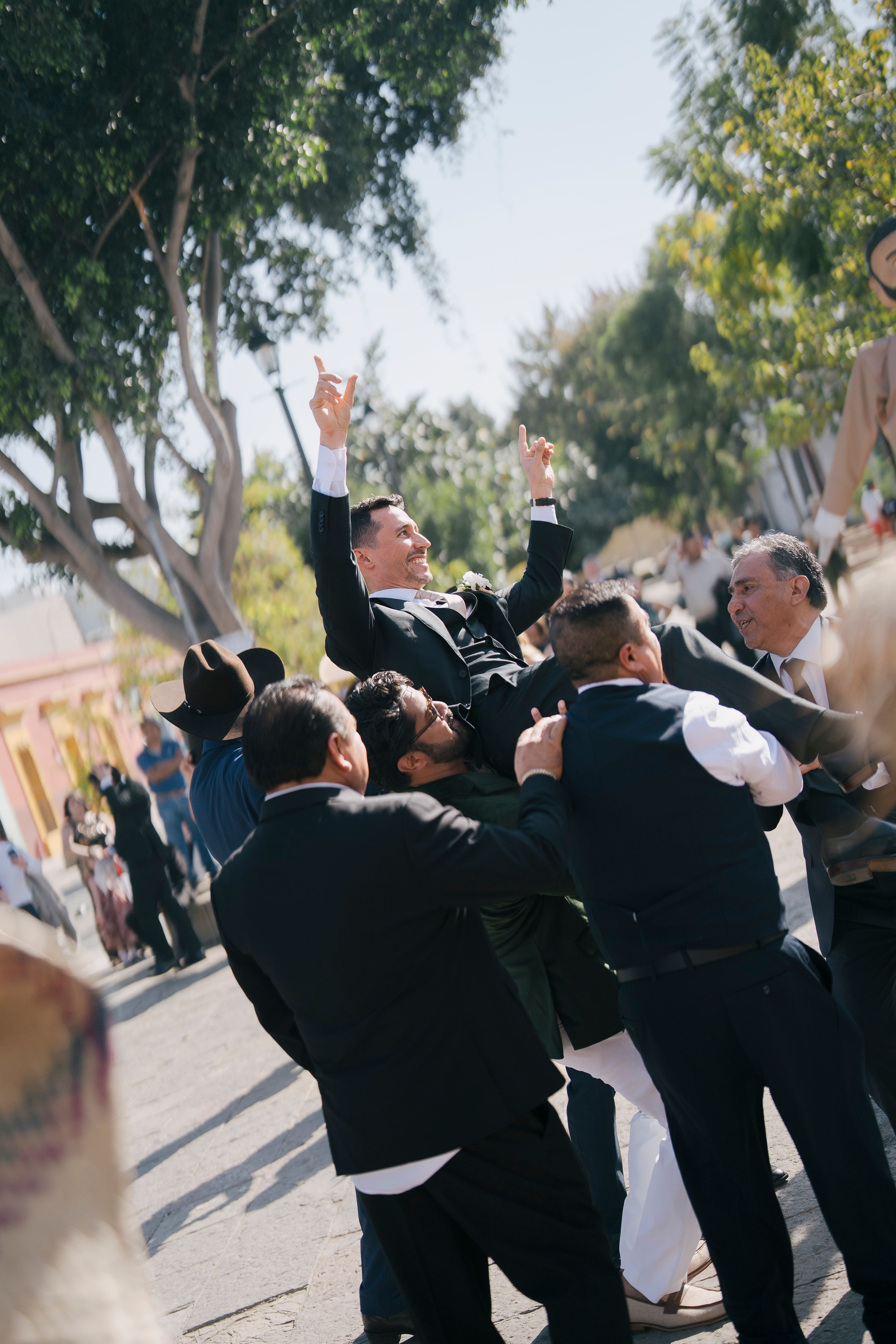 Groom being lifted by groomsmen outdoors at wedding celebration, raising hands joyfully with trees in sunny background.