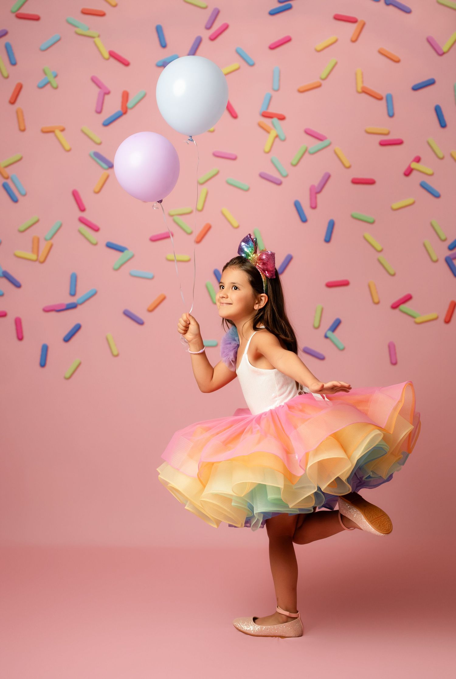A young girl in a rainbow tutu twirls with balloons on a pink background with colorful sprinkles falling around her.