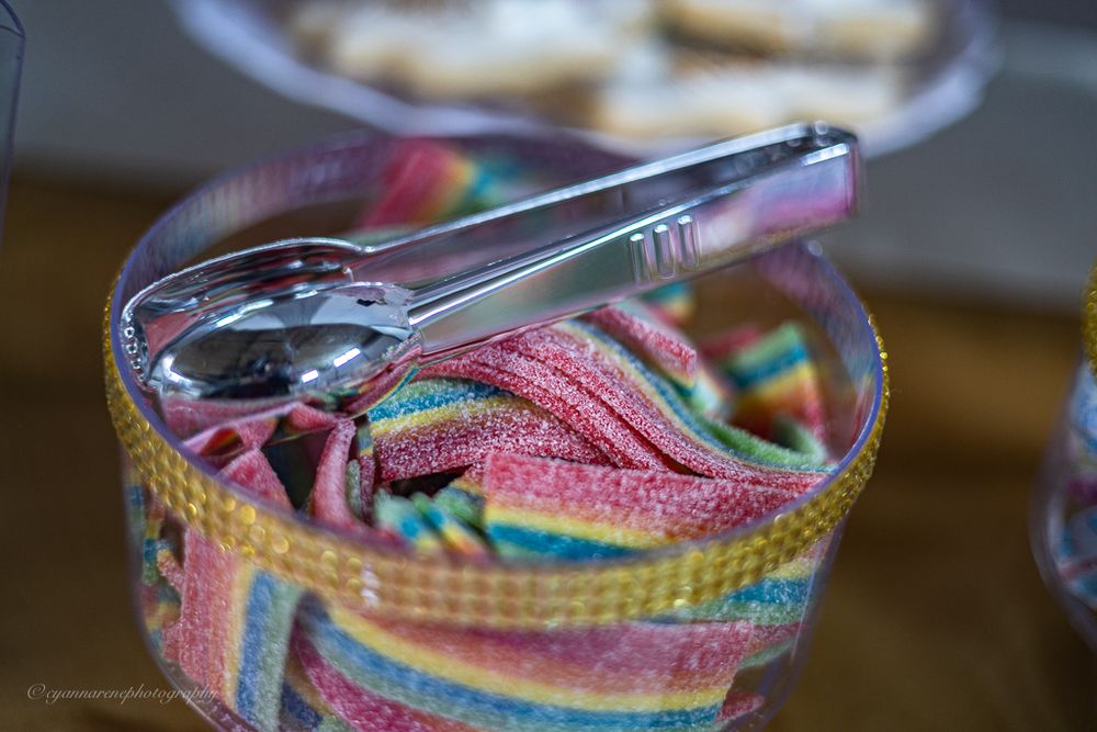 Glass jar filled with rainbow striped sour gummy candy with silver tongs at a party.