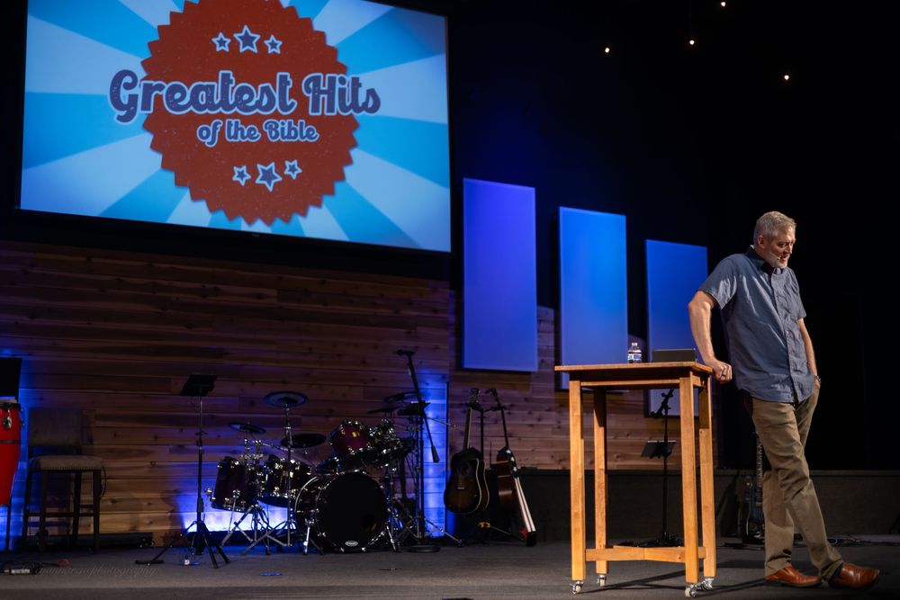 Pastor speaks at wooden podium on church stage with Greatest Hits of the Bible presentation screen behind him.