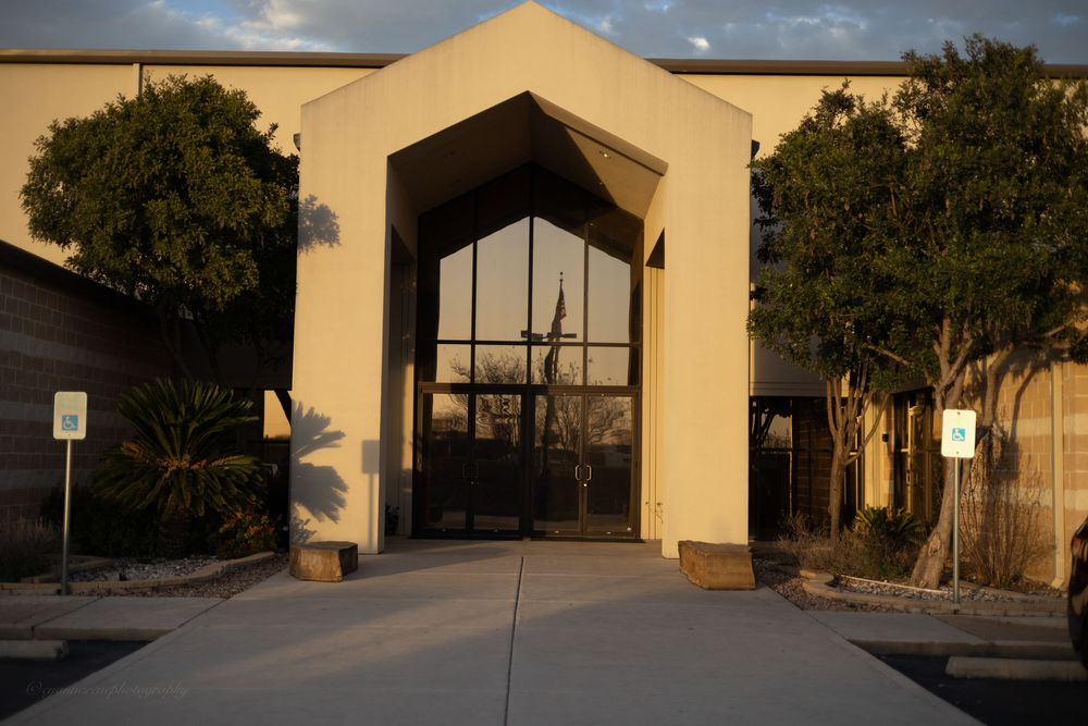 Exterior entrance of a tan stucco building with arched glass doors, surrounded by desert landscaping at sunset.