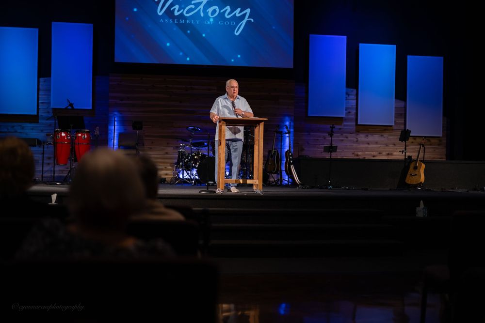 Pastor speaking at a wooden podium on a church stage with Victory sign and blue stage lighting.