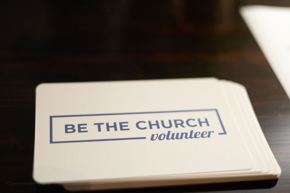 Stack of white "Be The Church Volunteer" cards on a dark wooden table.