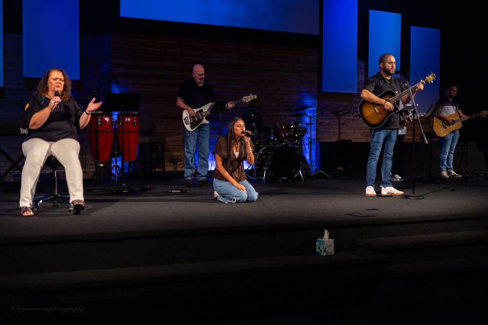 Band performs worship music on church stage with singers, guitarist, and drummer under blue stage lighting.