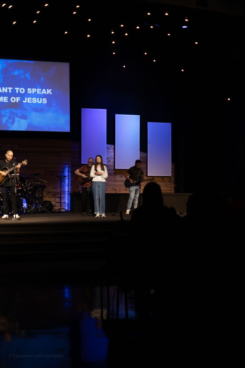 Christian worship band performing on a dimly lit church stage with projected lyrics reading the name of Jesus.