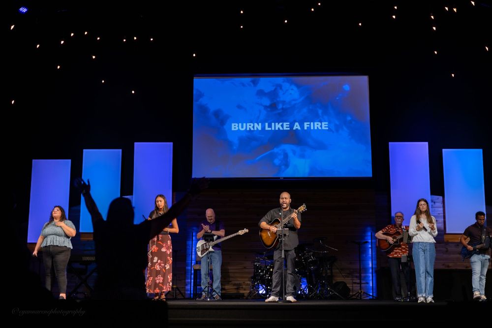 Worship band performs on stage with blue lighting and 'Burn Like A Fire' lyrics displayed on screen.
