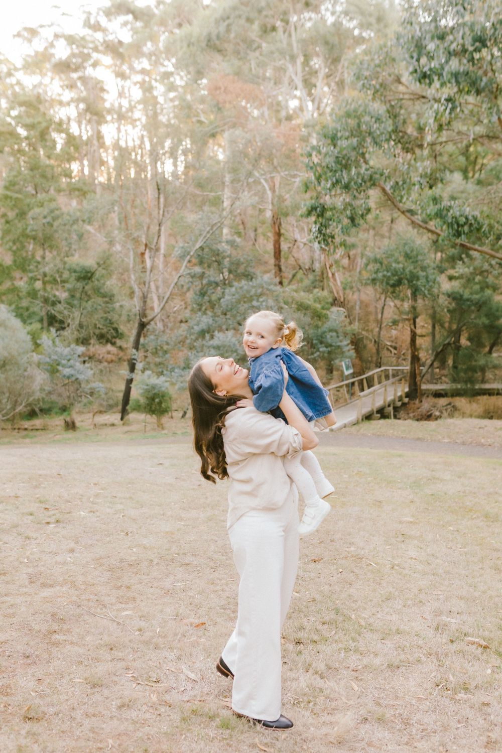 A woman in white lifts a laughing toddler in a denim jacket outdoors in a lush green park setting.