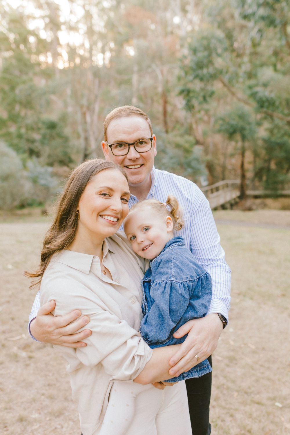 Family of three smiling outdoors, parents holding young girl in denim dress, surrounded by trees.