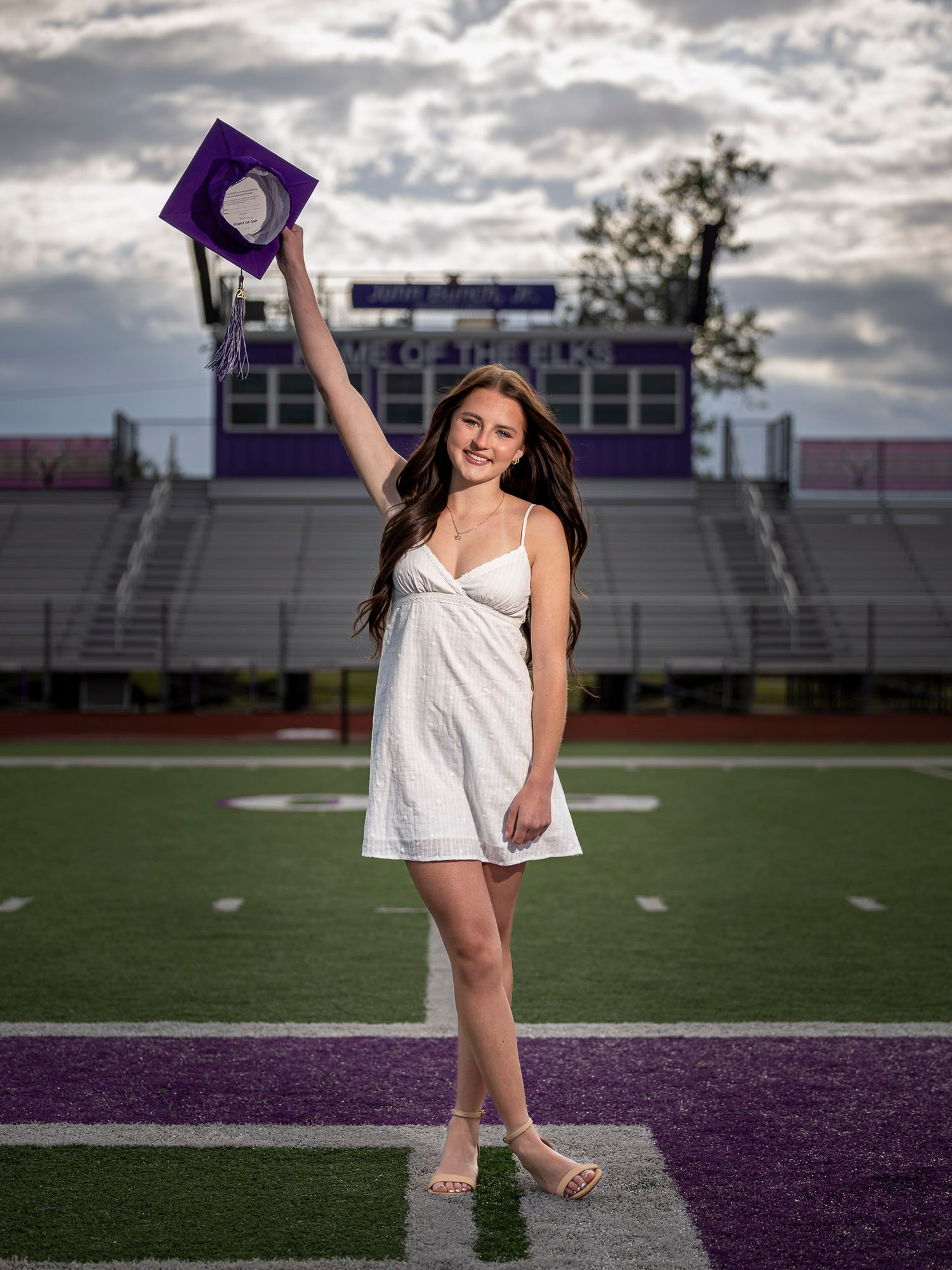 Young woman in white dress holds purple graduation cap aloft on a football field with purple stadium bleachers.
