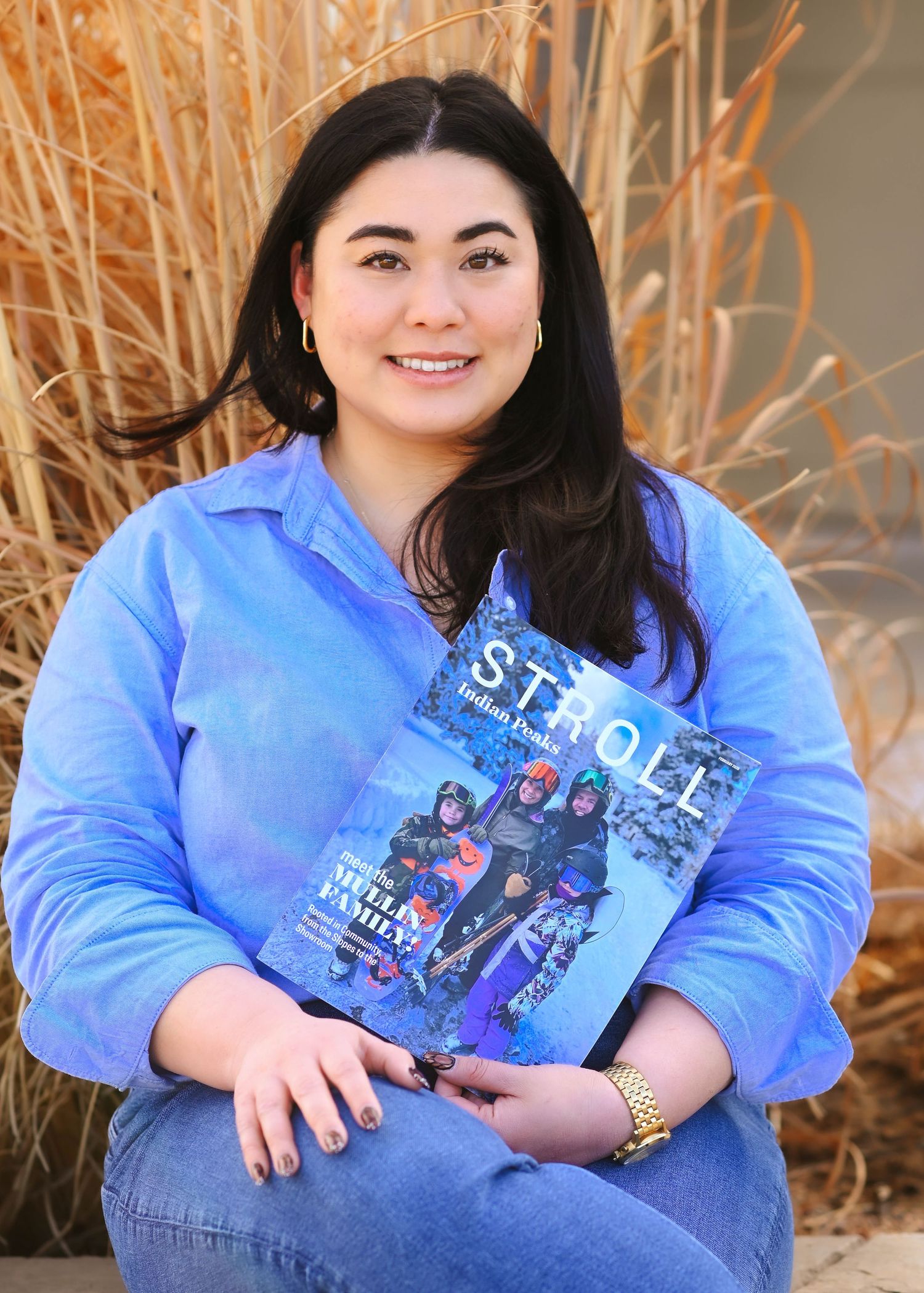 Woman in blue shirt smiling while holding a Stroll Indian Hills magazine, seated outdoors.