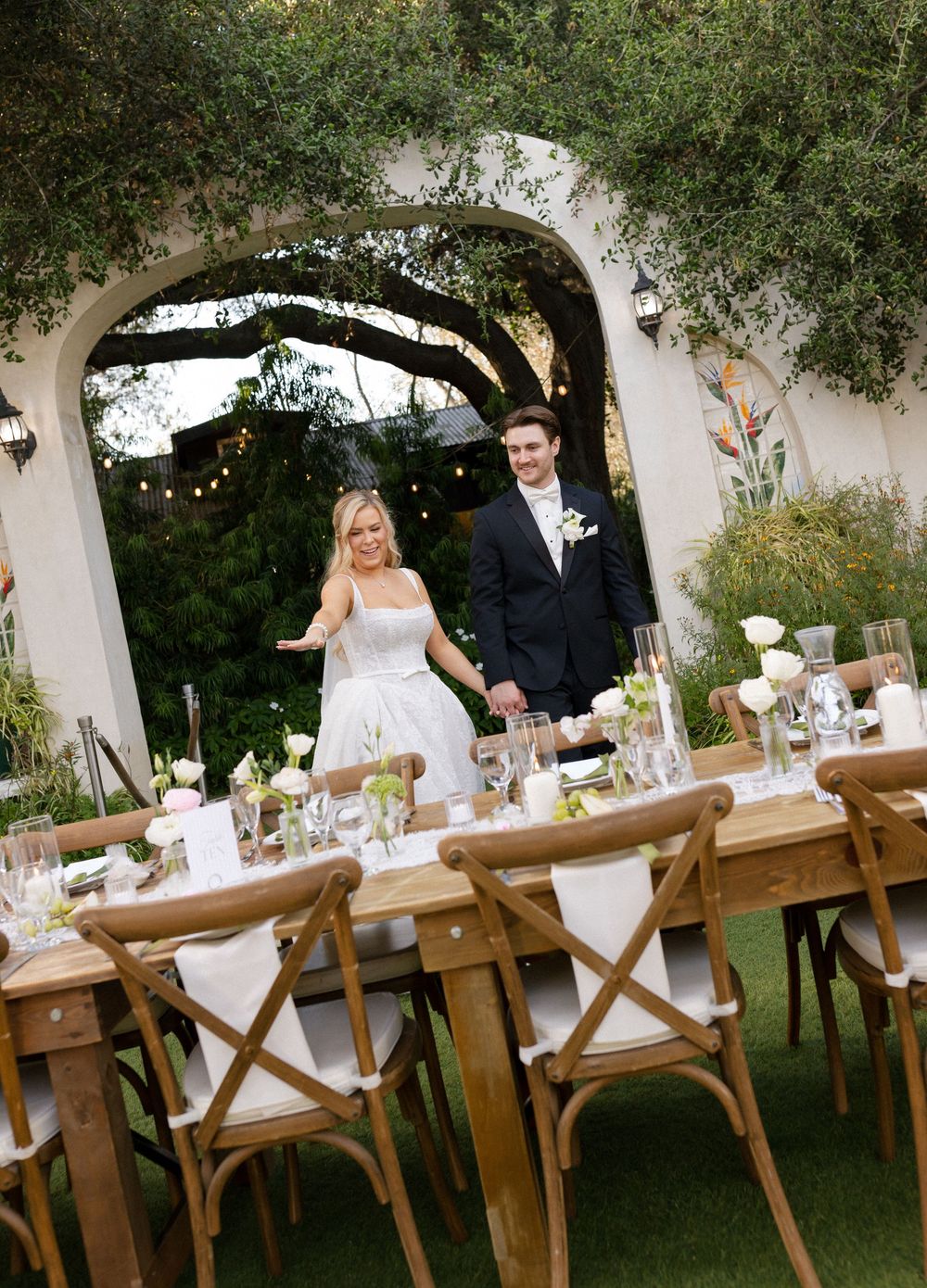 Smiling bride and groom entering elegant outdoor wedding reception with rustic wooden tables.