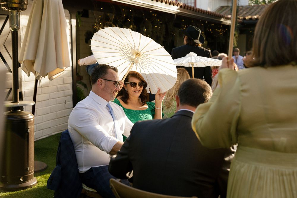 Wedding guests socializing outdoors with white parasol at sunny garden reception venue.