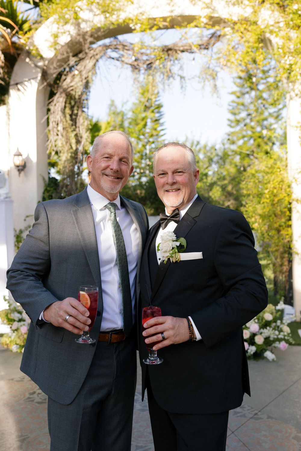 Two men in suits smiling and holding drinks in front of floral wedding arch.