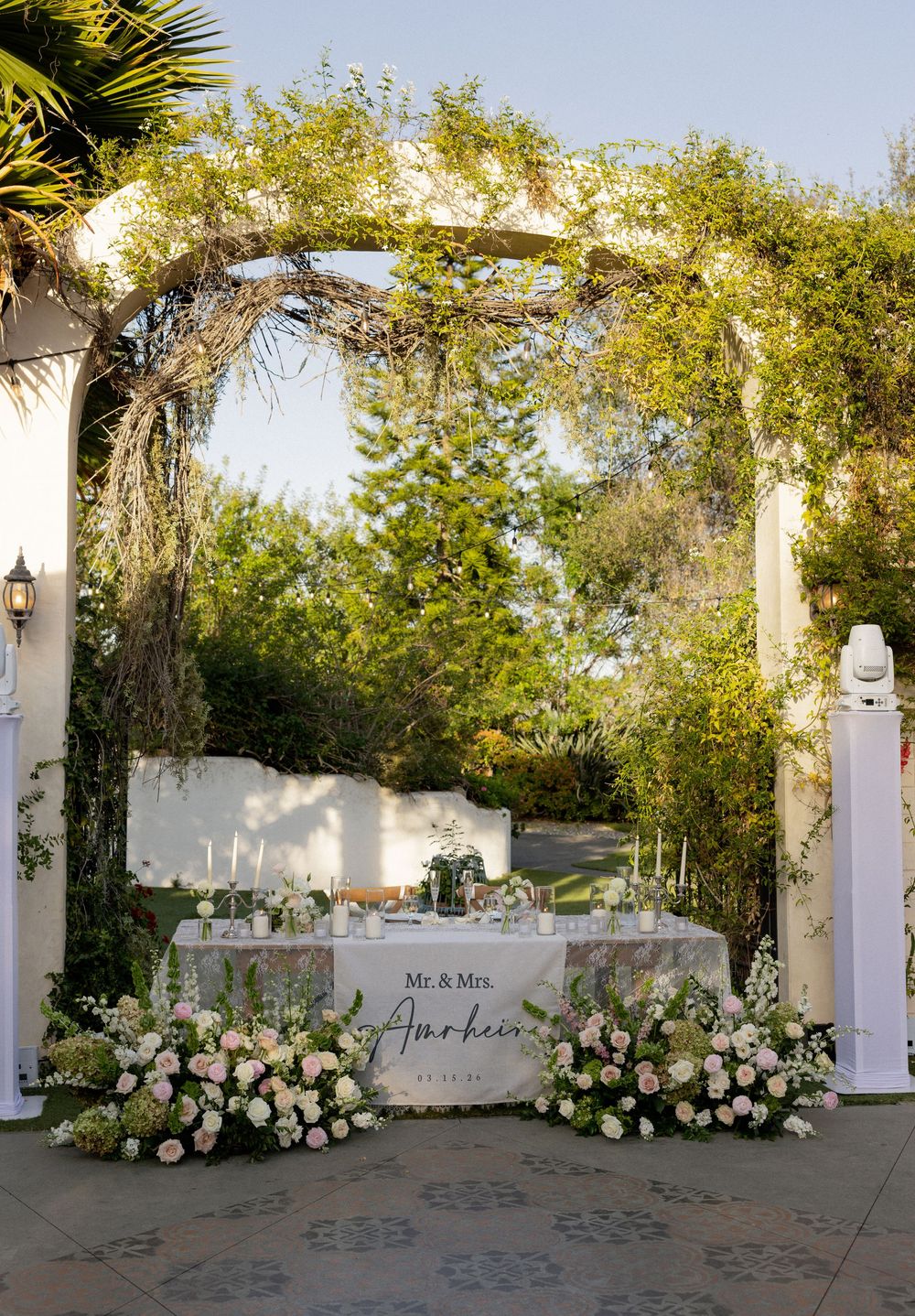 Mr. and Mrs. sweetheart table adorned with pink roses under a floral pergola at outdoor wedding.