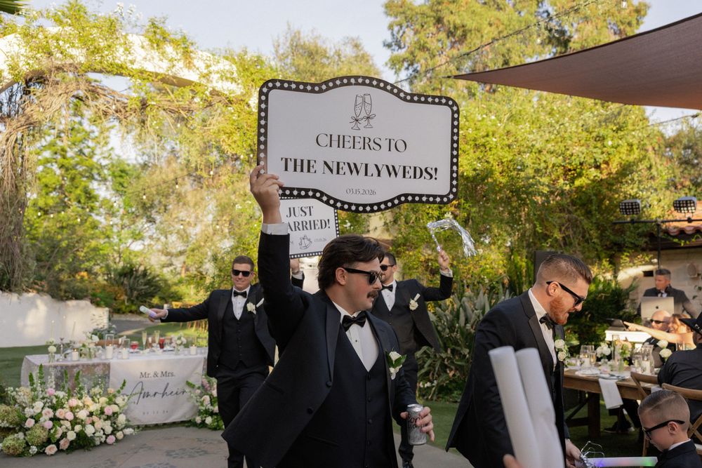 Groomsman holds a cheers to the newlyweds sign at an elegant outdoor wedding reception celebration.