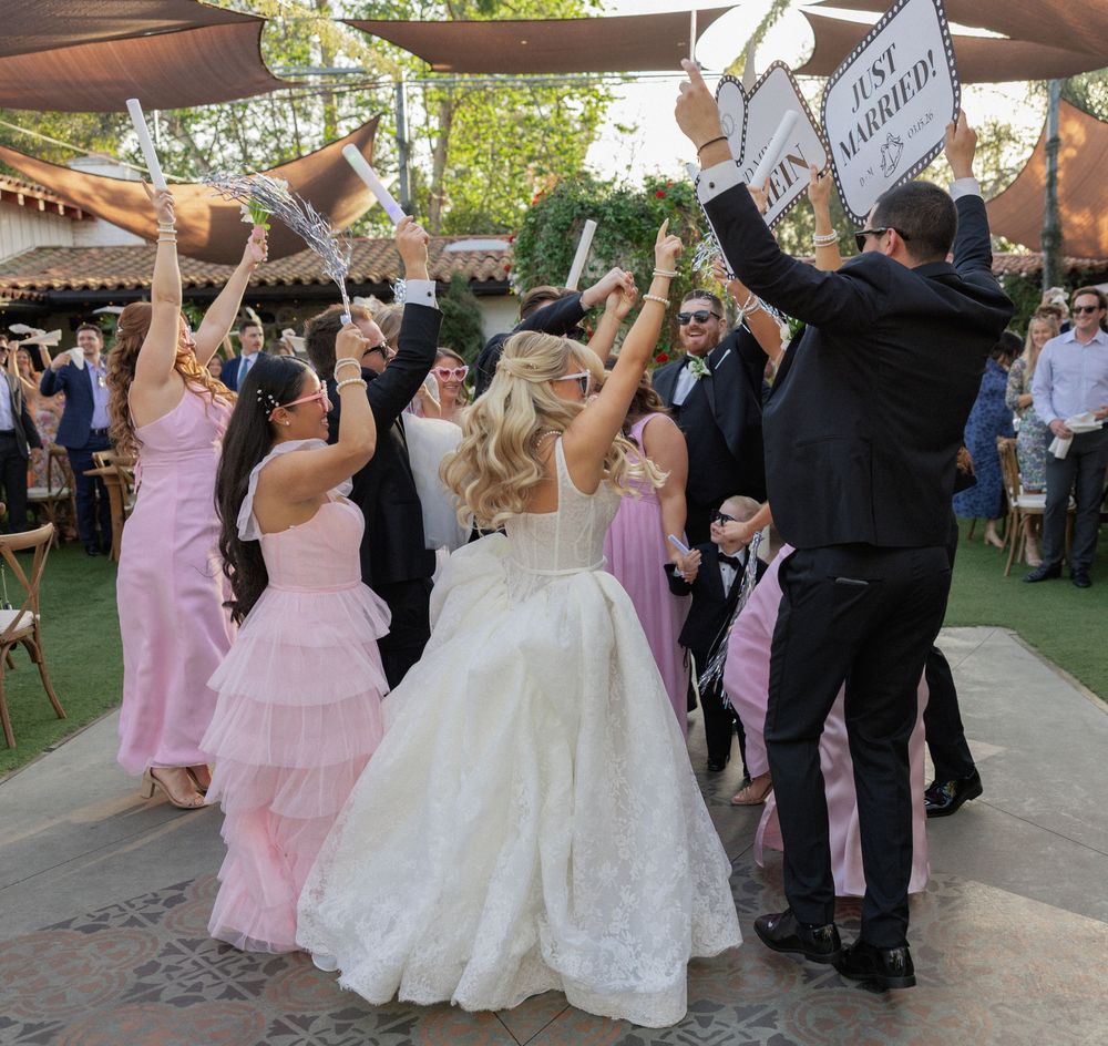Wedding party cheering with raised signs as bride in white gown celebrates with guests at outdoor garden reception.