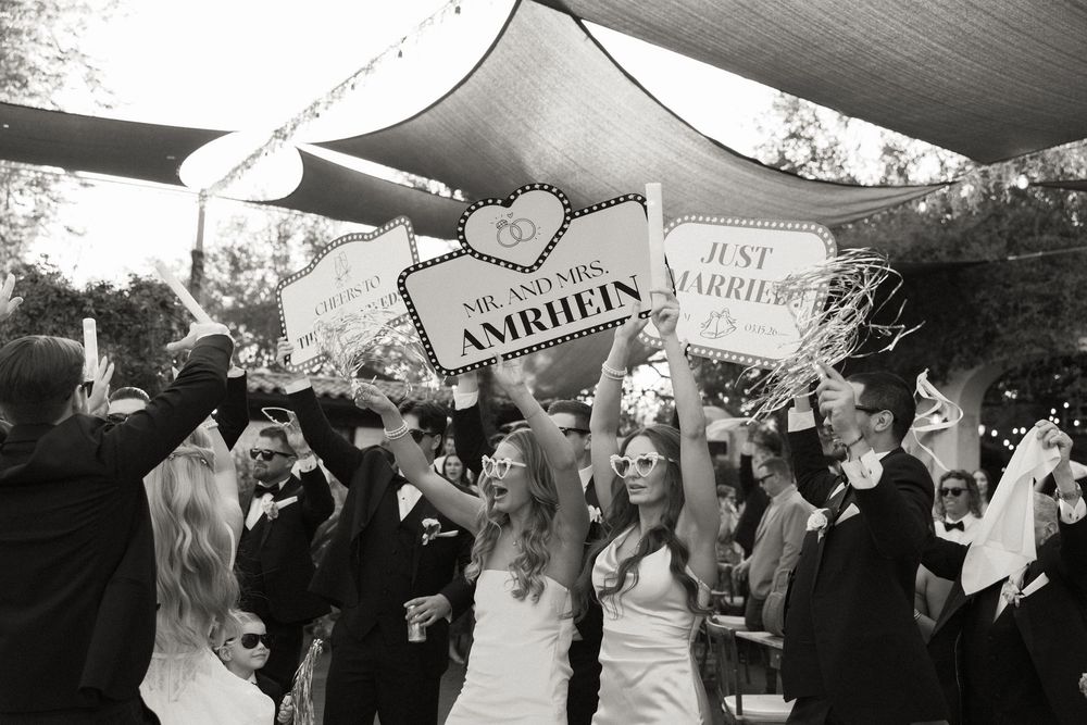 Bride and groom celebrate with guests holding 'Just Married' and 'Mr. and Mrs. Amrhein' signs outdoors.