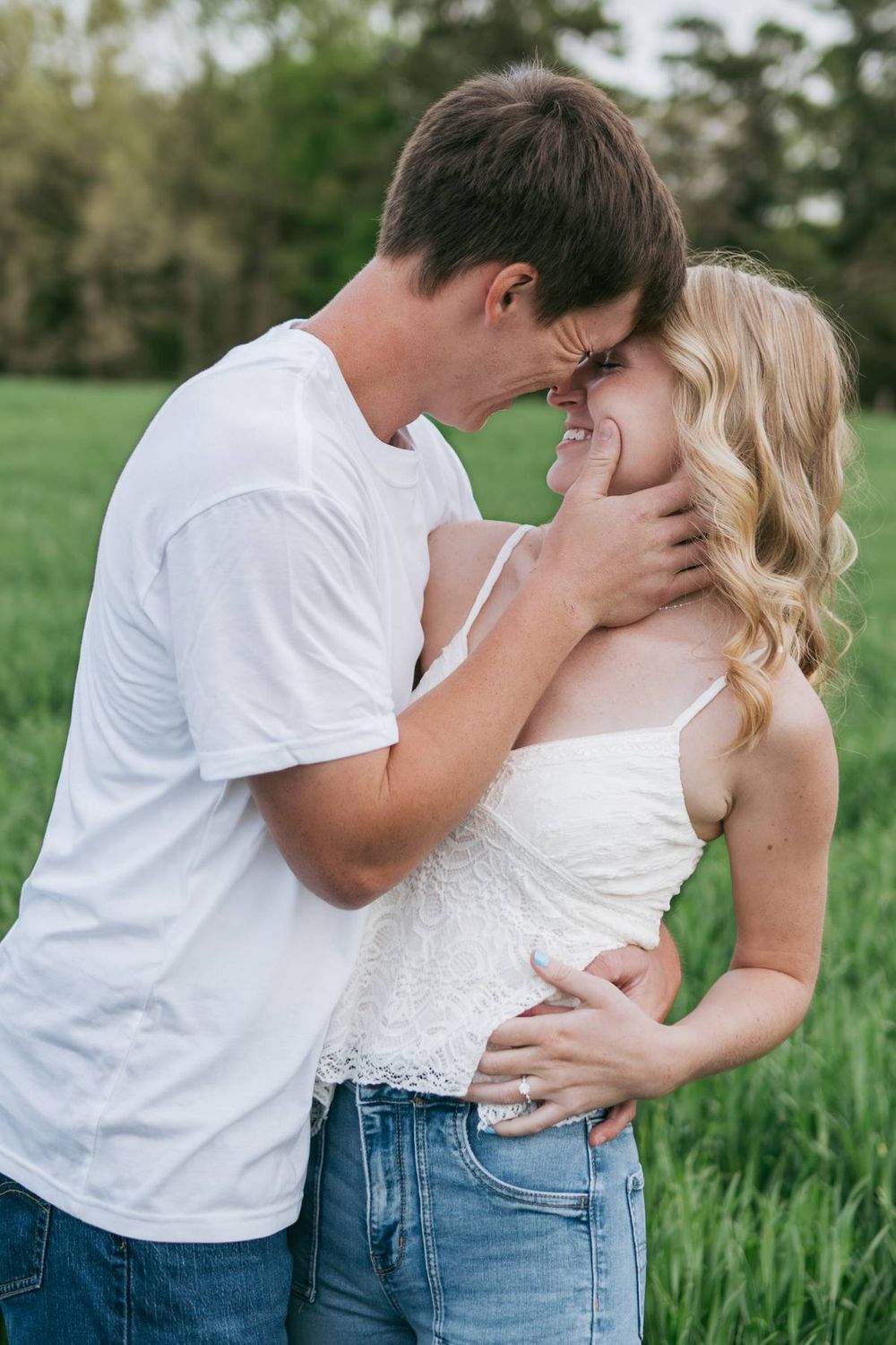Happy engaged couple nearly kissing in a green field, woman smiling in white top and jeans, engagement photographer Lexington NC
